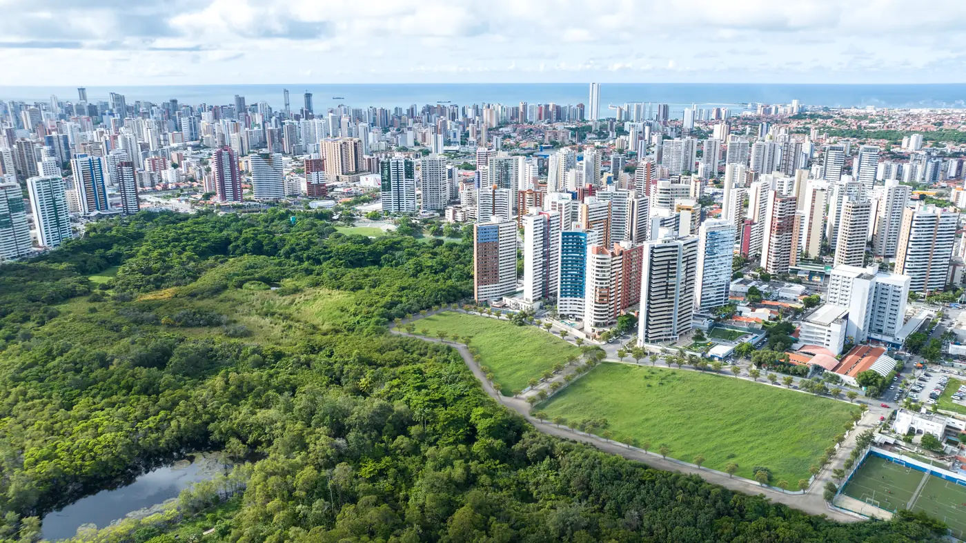 Foto aérea do terreno ainda vazio na borda do Parque do Cocó, cercado por edifícios já construídos, com o mar ao fundo